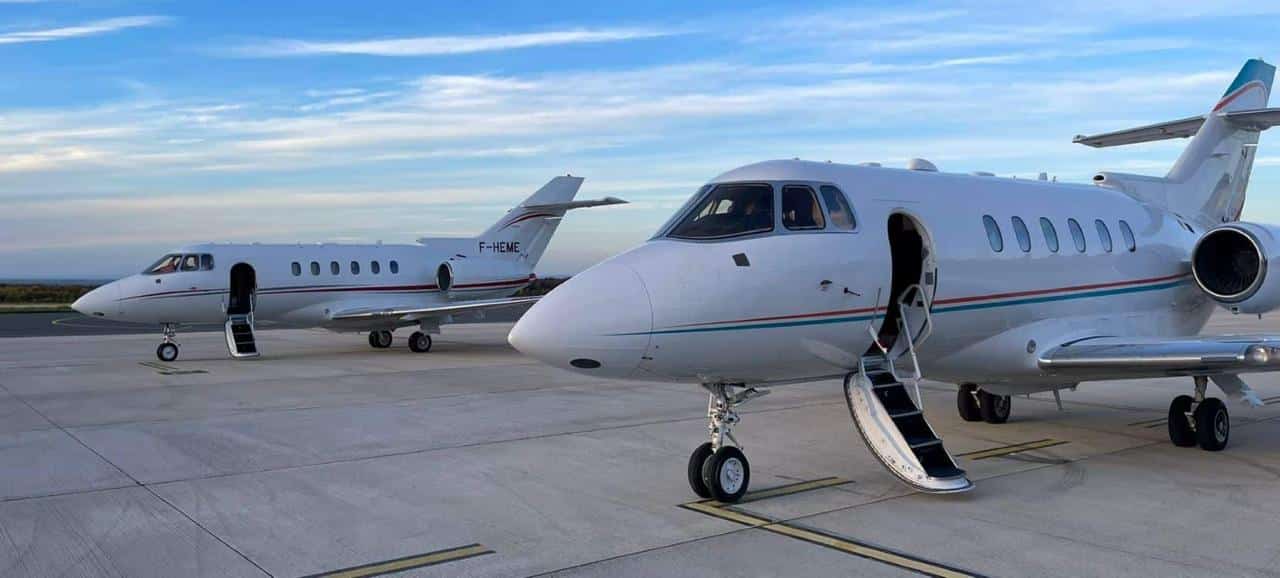 Deux jets privés blancs stationnés sur un tarmac d’aéroport, avec leurs portes ouvertes et escaliers déployés, sous un ciel légèrement nuageux.